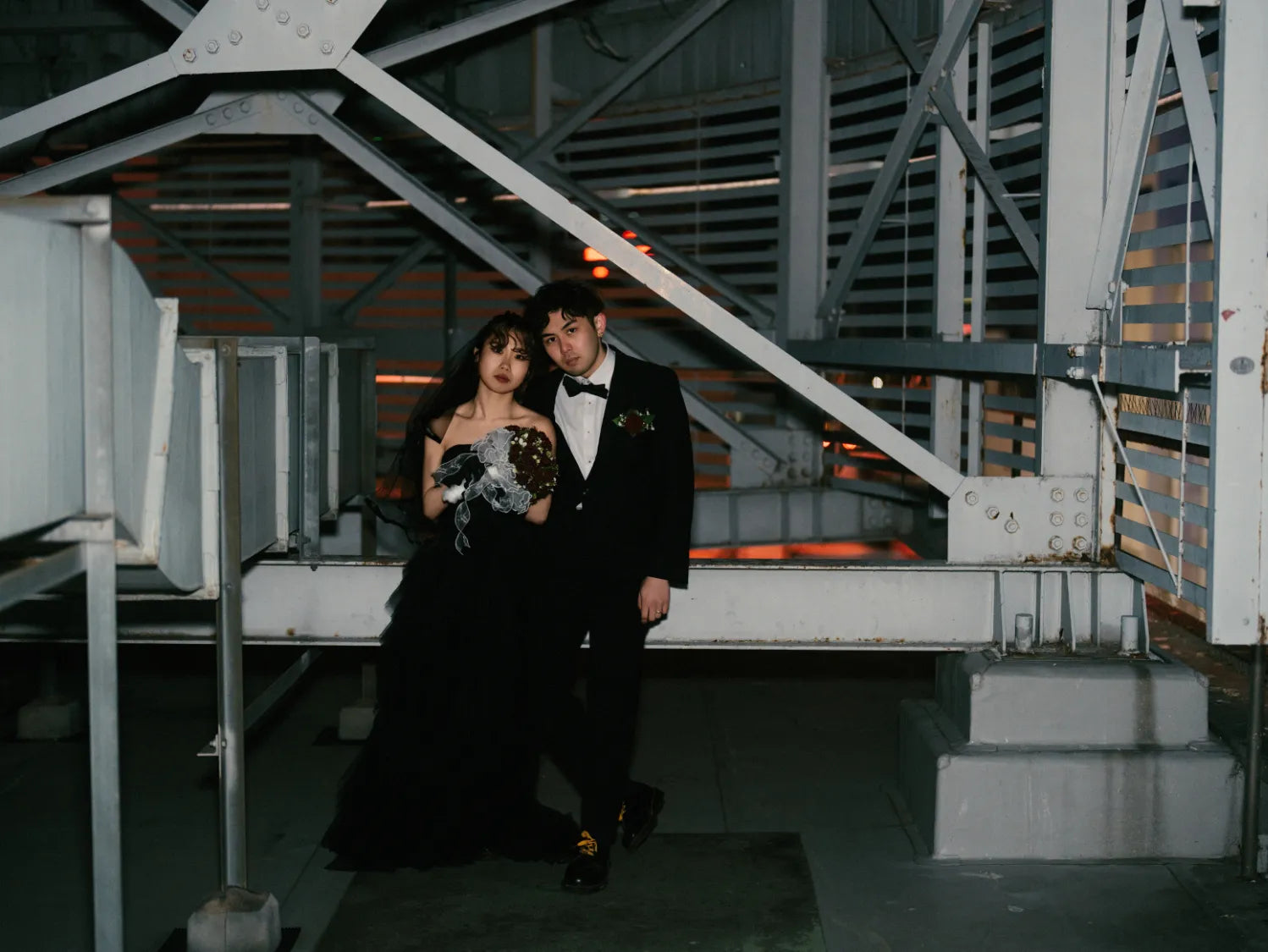 工場のような夜の屋上構造物の中で寄り添う黒ドレスとタキシードのカップル。夜の雰囲気と都会的なムードが魅力のウェディングフォト。仙台フォトスタジオQnNe(キュンネ)