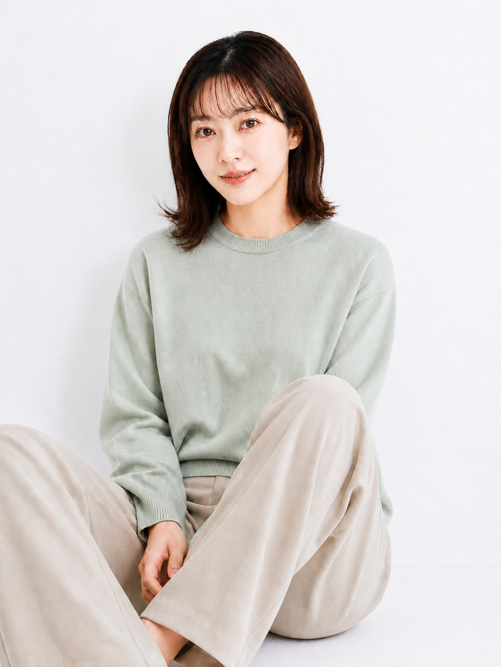 白いスタジオでセージ色のニットとグレージュのパンツを着て座る日本人女性のポートレート