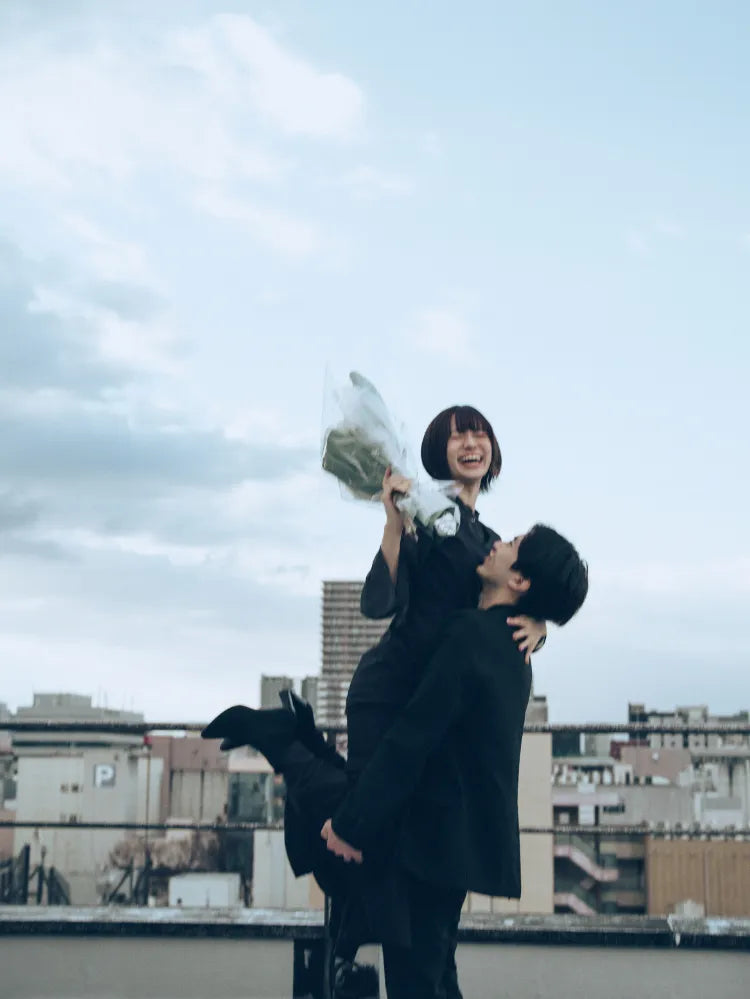 仙台のビル屋上で花束を手に笑顔を見せる女性を抱き上げる男性。プロ撮影によるナチュラルで感動的なカップル写真。仙台フォトスタジオQnNe(キュンネ)